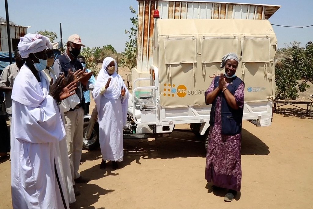 تقف ناتاليا كانيم مبتسمة وتصفق أمام عيادة متنقلة تابعة UNFPA في السودان، بينما يصفق أفراد آخرون من المجتمع المحلي أيضاً.
