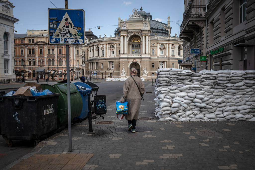 Une femme marche dans une rue en Ukraine, en passant devant un grand tas de sacs de sable, avec l'Opéra d'Odessa en arrière-plan.