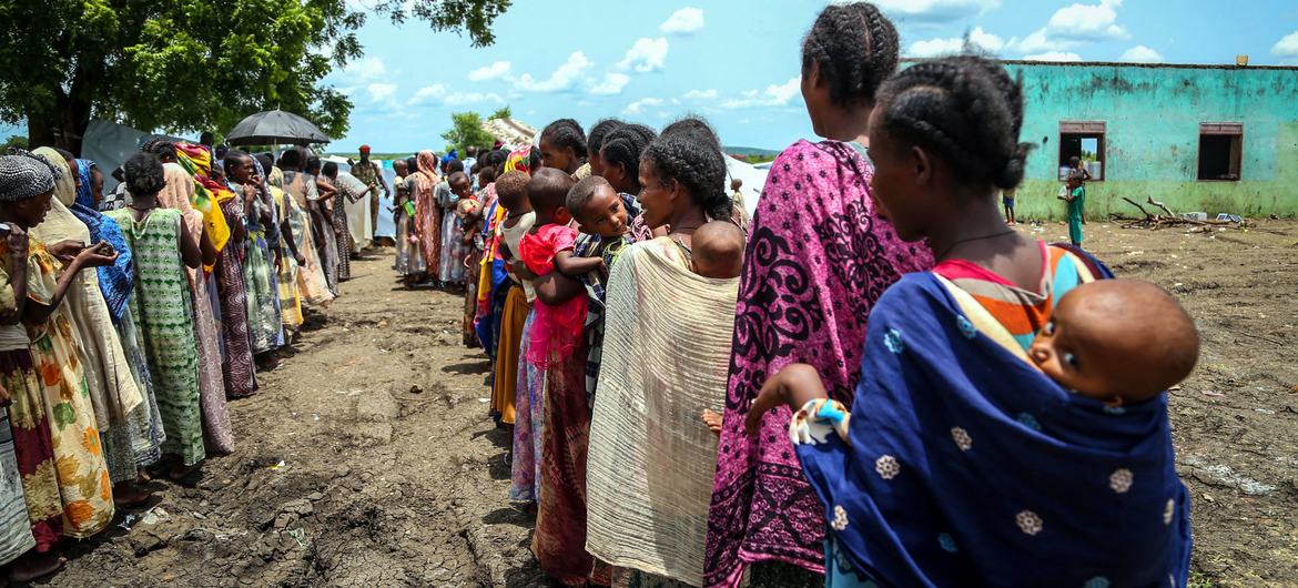 Uma longa fila de mulheres e crianças, algumas carregando bebês, aguarda ao ar livre em uma área rural sob um céu claro e ensolarado.