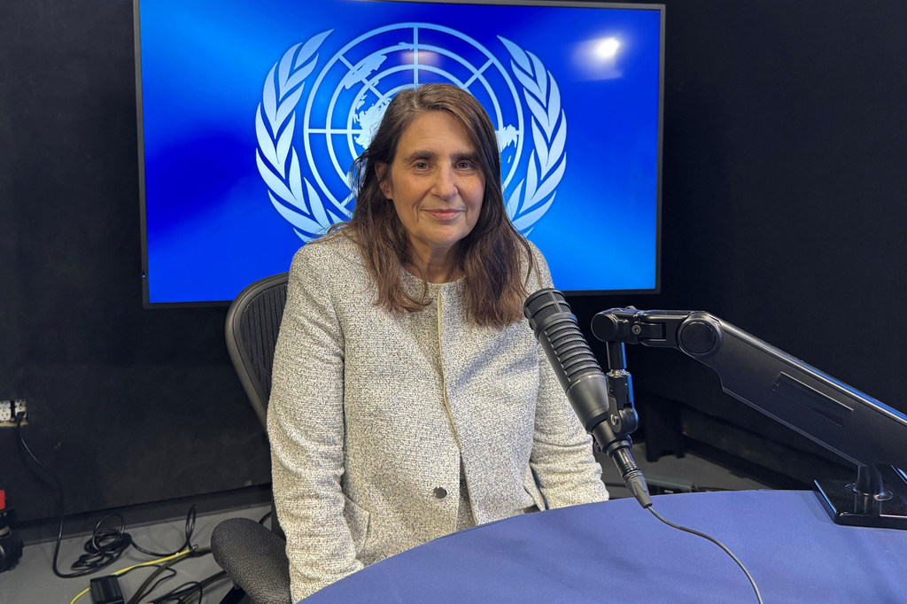 La Sra. Shari Spiegel está sentada en una mesa con un micrófono frente a ella, hablando durante una entrevista Noticias ONU , con el logotipo de la ONU visible en una pantalla detrás de ella.