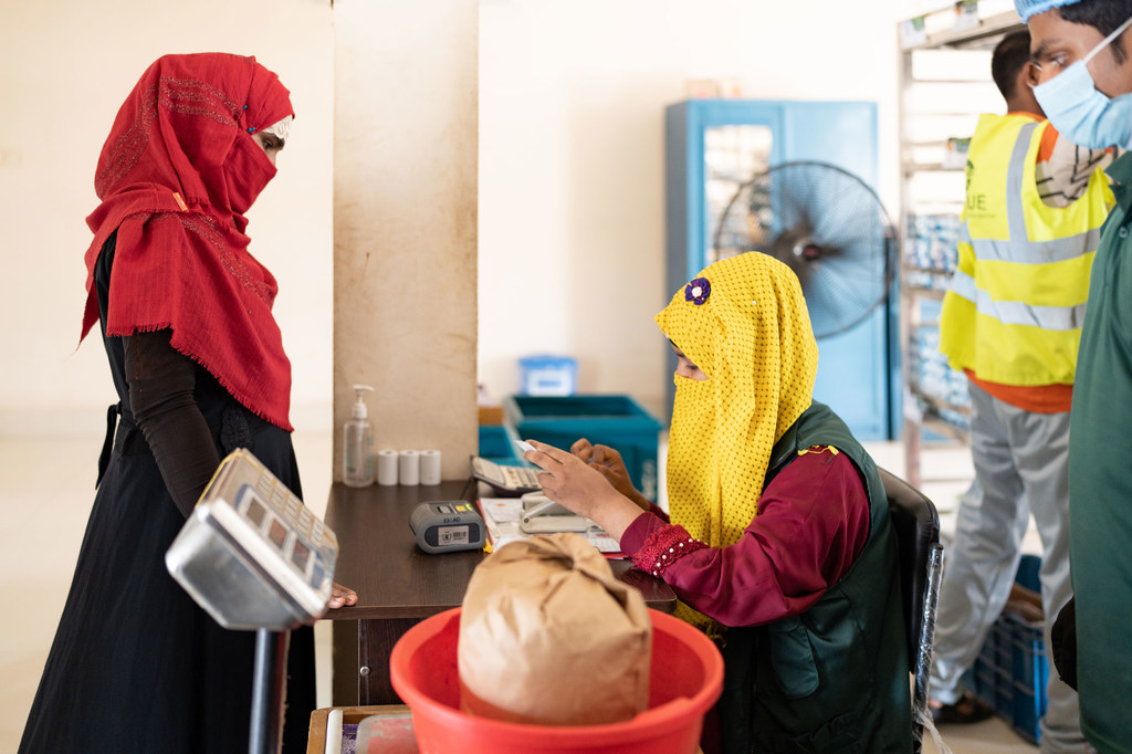 Mfanyakazi wa misaada WFP akiwa amevaa kitambaa cha manjano na fulana akimgawia mgao wa chakula mwanamke mkimbizi wa Rohingya aliyevaa kitambaa chekundu.