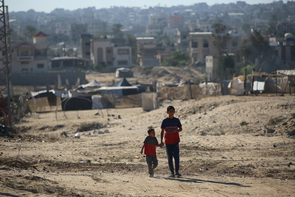 صبيان فلسطينيان يسيران يداً بيد على طول طريق ترابي في منطقة مليئة بالأنقاض، مع وجود مبانٍ متضررة في الخلفية في خان يونس، قطاع غزة.