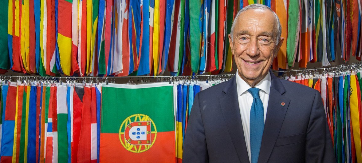 Marcelo Rebelo de Sousa, Presidente de Portugal, sorri para um retrato formal em frente a uma parede de bandeiras internacionais, incluindo a bandeira portuguesa.