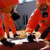 Сотрудники экстренных служб в оранжевых защитных костюмах и противогазах отрабатывают медицинские процедуры на тренировочном манекене.