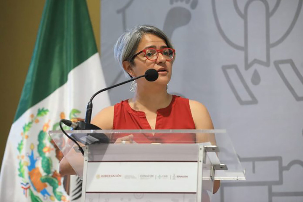 La mujer Karla Quintana habla desde un podio con una bandera mexicana detrás de ella.