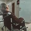Пожилой человек с седыми волосами сидит в инвалидном кресле в больничном коридоре.