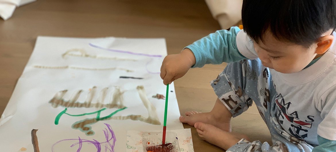 Um menino asiático de três anos, de pijama, está pintando em uma grande folha de papel sobre um piso de madeira.