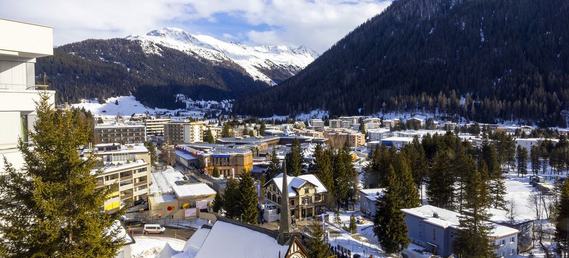 Uma vista panorâmica da cidade suíça de Davos, uma estância de esqui, no inverno, com edifícios cobertos de neve aninhados entre pinheiros na base de uma cordilheira florestada.