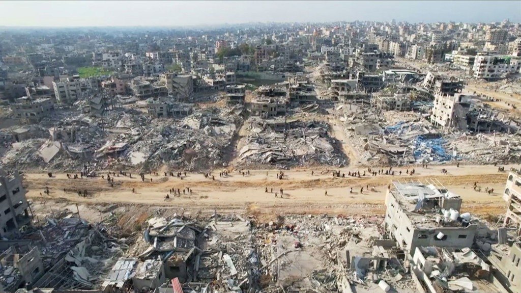 صورة جوية لمدينة في غزة تُظهر دمارًا واسع النطاق مع أنقاض ومباني متضررة، بينما يمشي الناس على طريق ترابي في المقدمة.