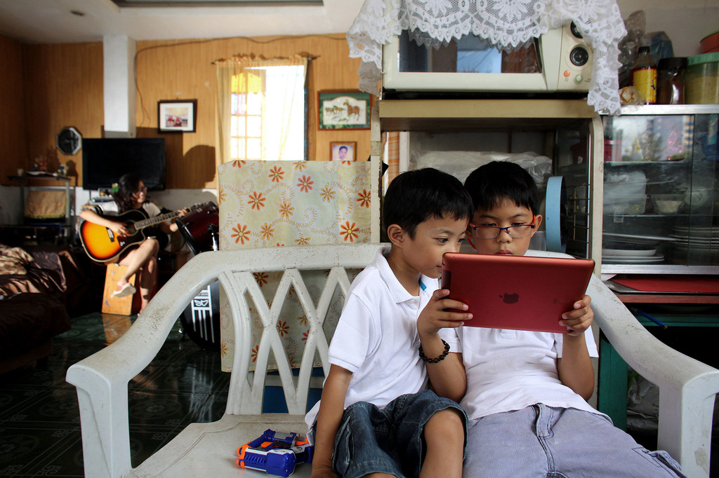 Dans une maison à Cebu, aux Philippines, deux jeunes garçons asiatiques regardent ensemble une tablette rouge. En arrière-plan, une femme joue de la guitare acoustique.