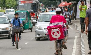 A food delivery cyclist wearing a pink delivery bag with a panda logo rides through busy urban traffic in Bangladesh.