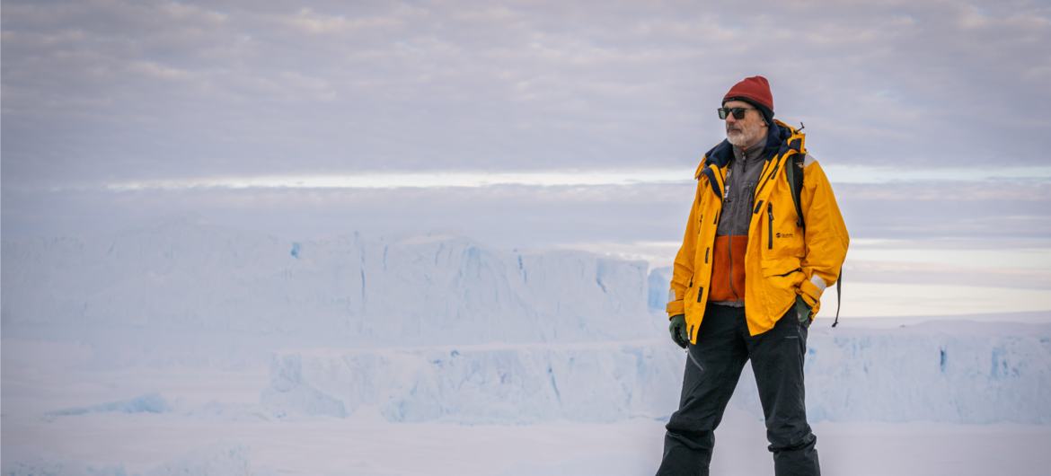 Pesquisador brasileiro em pé sobre um afloramento rochoso na Antártica, com uma vasta geleira e formações de gelo ao fundo.