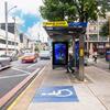 Vista de una parada de autobús moderna en una calle urbana, con un anuncio de JCDecaux, un símbolo azul de acceso para personas con discapacidad en el suelo y vehículos circulando por la carretera.