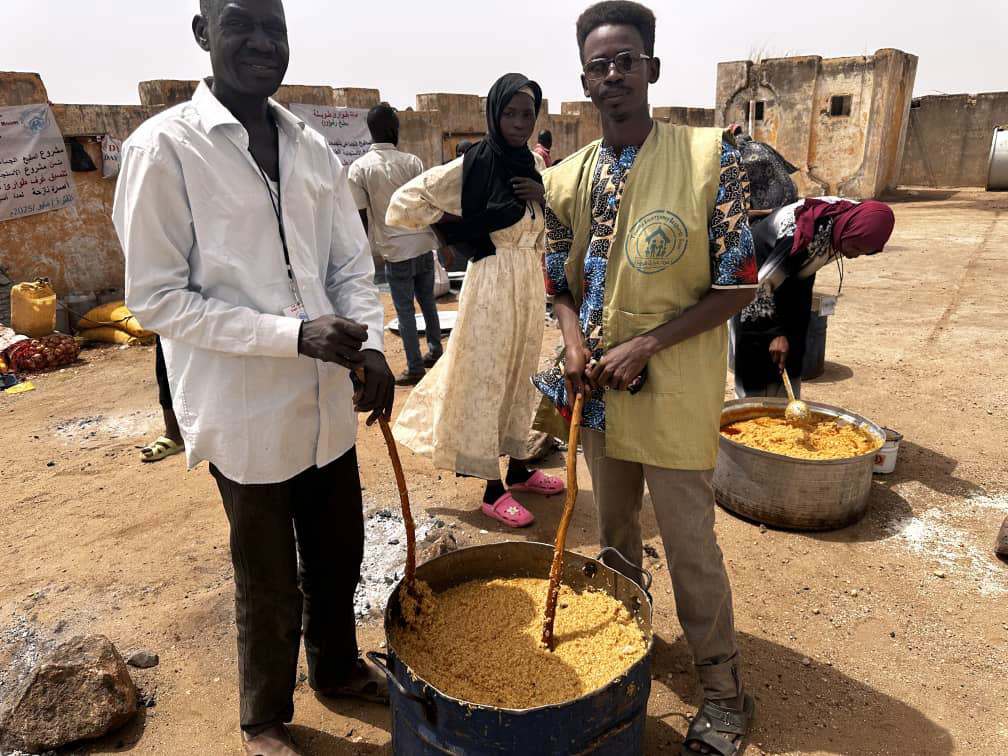 رجال وامرأة سودانيون يقفون بجوار أوانٍ كبيرة من الطعام في مطبخ مجتمعي في دارفور، السودان، كجزء من مبادرة الإغاثة "طويلة".