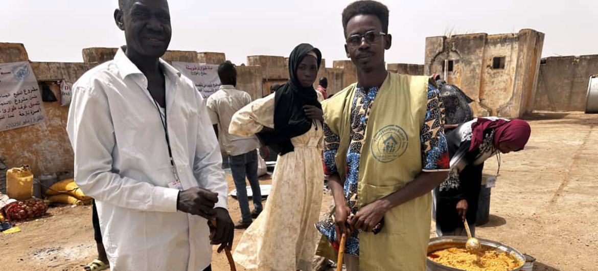 Homens e uma mulher sudaneses posam ao lado de grandes panelas de comida em uma cozinha comunitária em Darfur, Sudão, como parte da iniciativa de ajuda humanitária Taweela.
