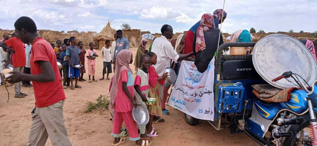 توزيع مساعدات مجتمعية في دارفور، السودان، حيث يقوم شاب بتوزيع الطعام على الأطفال والبالغين المتجمعين حول شاحنة. وفي الخلفية، يظهر كوخ مسقوف بالقش وقدر طهي كبير.