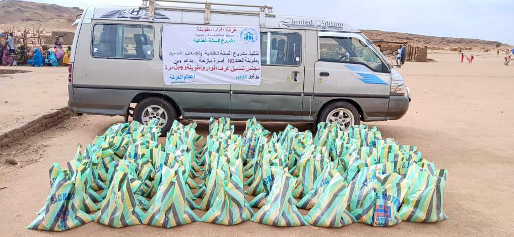 شاحنة تحمل لافتة عربية تقف خلف العديد من أكياس المساعدات الغذائية في منظر طبيعي قاحل ومغبر في منطقة الطويلة بشمال دارفور.