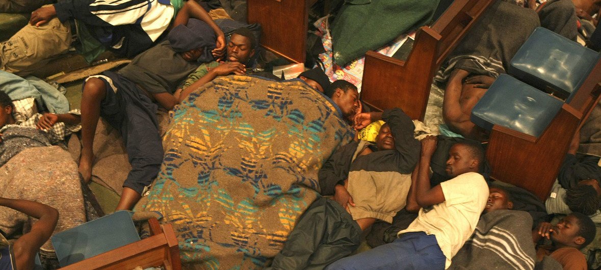 Homens dormem no chão da Igreja Metodista Central em Joanesburgo, usando os bancos como encosto de cabeça. A imagem mostra uma cena de aglomeração de refugiados em busca de abrigo.