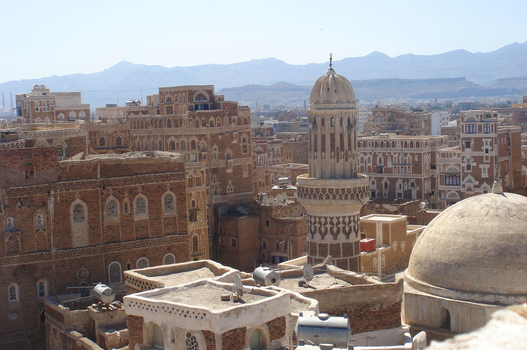 Vue panoramique de la vieille ville de Sana'a au Yémen, avec des bâtiments traditionnels en briques de terre et un minaret proéminent sur fond de montagnes.