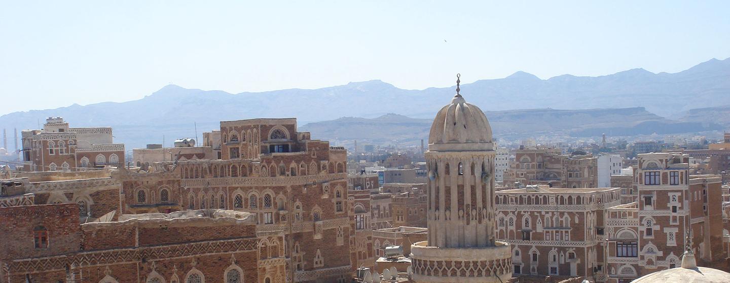 Vue panoramique de la vieille ville de Sana'a au Yémen, avec des bâtiments traditionnels en briques de terre et un minaret proéminent sur fond de montagnes.