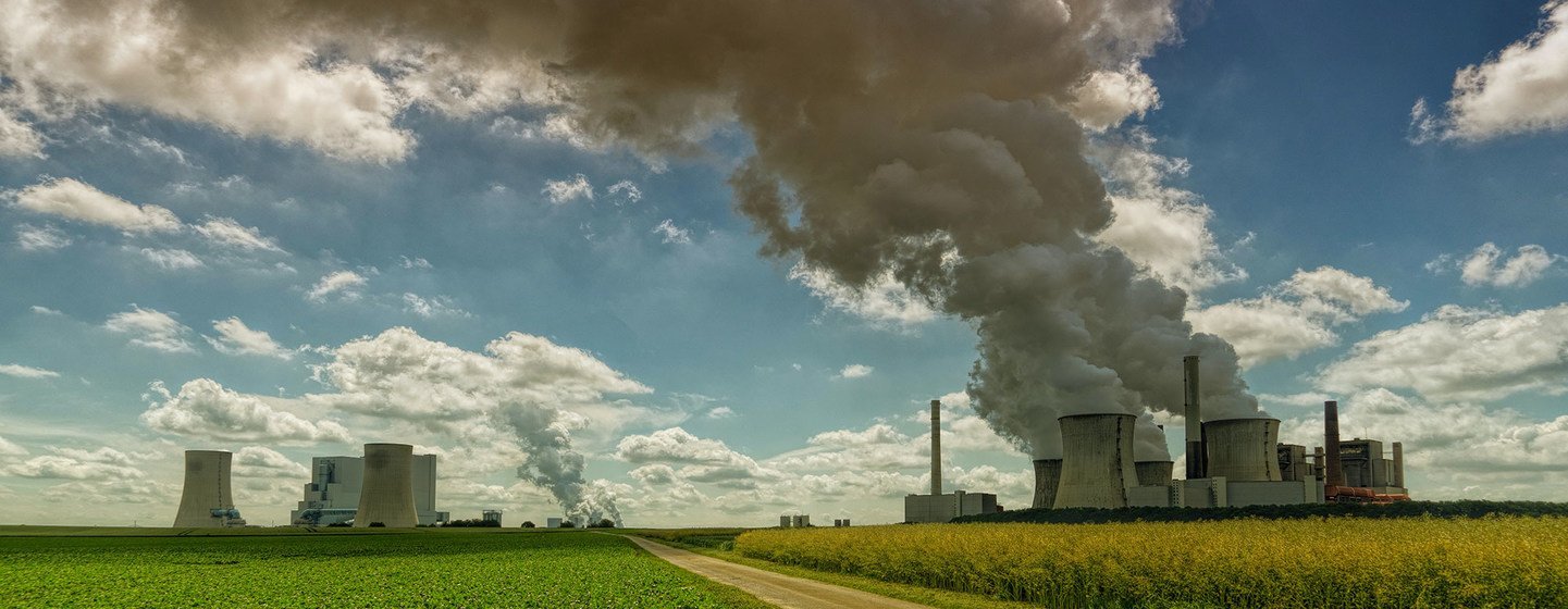 Un paysage rural parsemé de grandes centrales électriques industrielles dotées de tours de refroidissement et de cheminées crachant un épais panache de fumée dans un ciel partiellement nuageux.