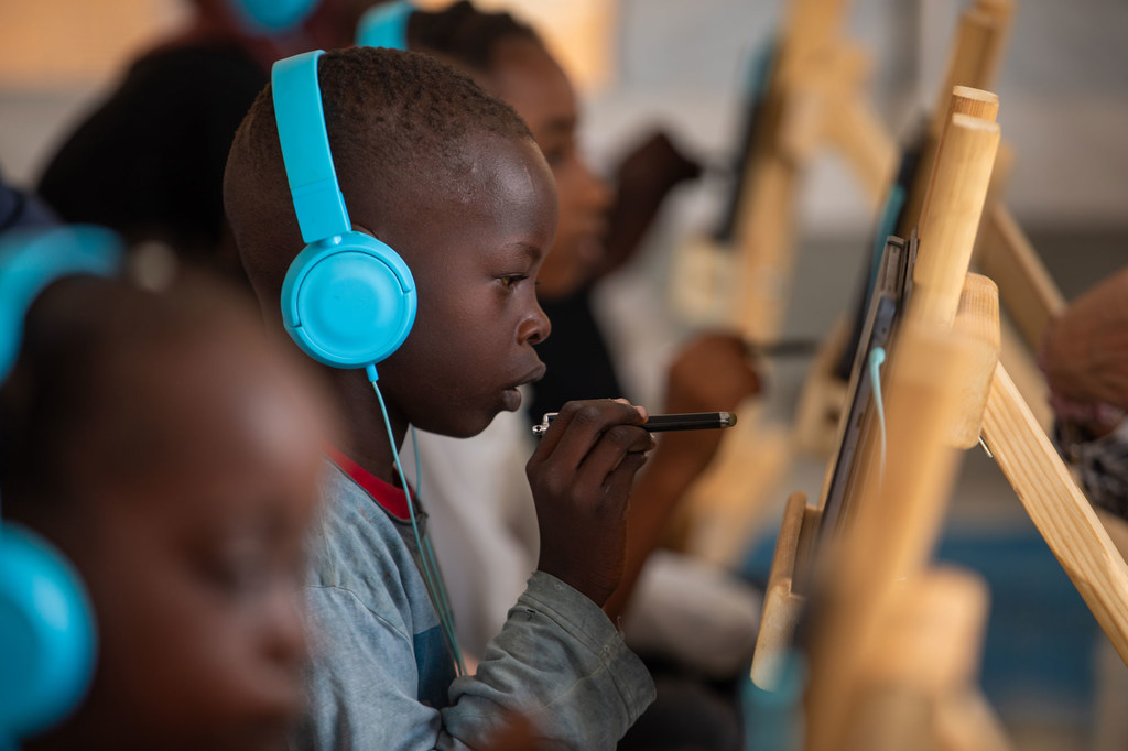Mvulana mdogo aliyevaa vipokea sauti vya masikioni vya bluu anatumia kompyuta kibao wakati wa kikao cha kujifunza mtandaoni katika eneo linalowafaa watoto UNICEF nchini Sudan.