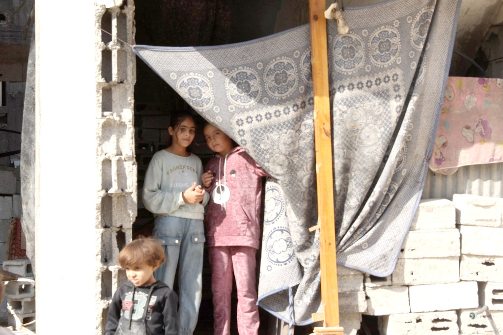 ثلاثة أطفال فلسطينيين يقفون أمام منزل مدمر في مدينة غزة، محاطين بالأنقاض والمأوى المؤقت.