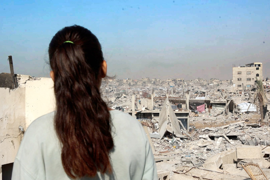 طفل فلسطيني يقف على سقف مبنى مدمر في غزة، ينظر إلى أفق المدينة المكون من الأنقاض والرمال.