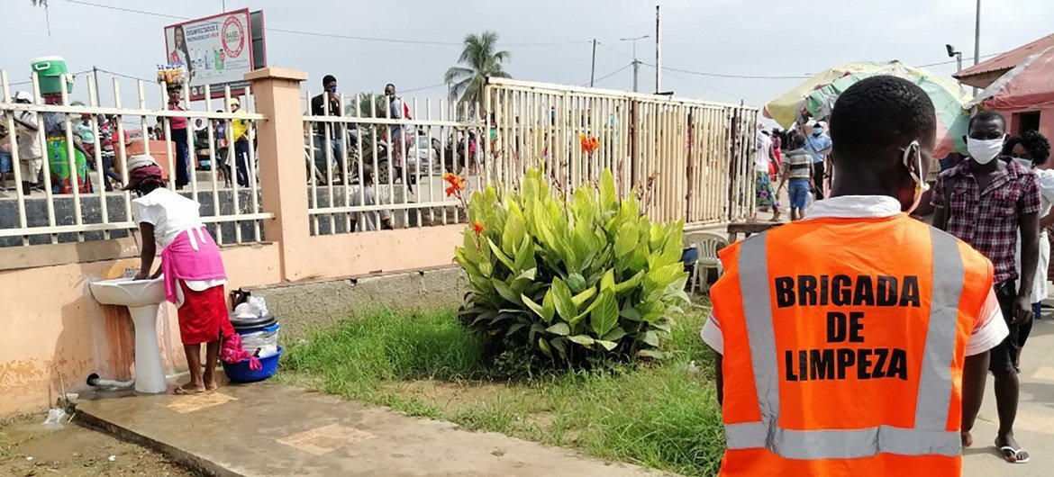 Um homem com um colete laranja da 'Brigada de Limpeza' observa uma mulher lavando as mãos em uma pia pública do lado de fora de um mercado em Luanda, Angola.