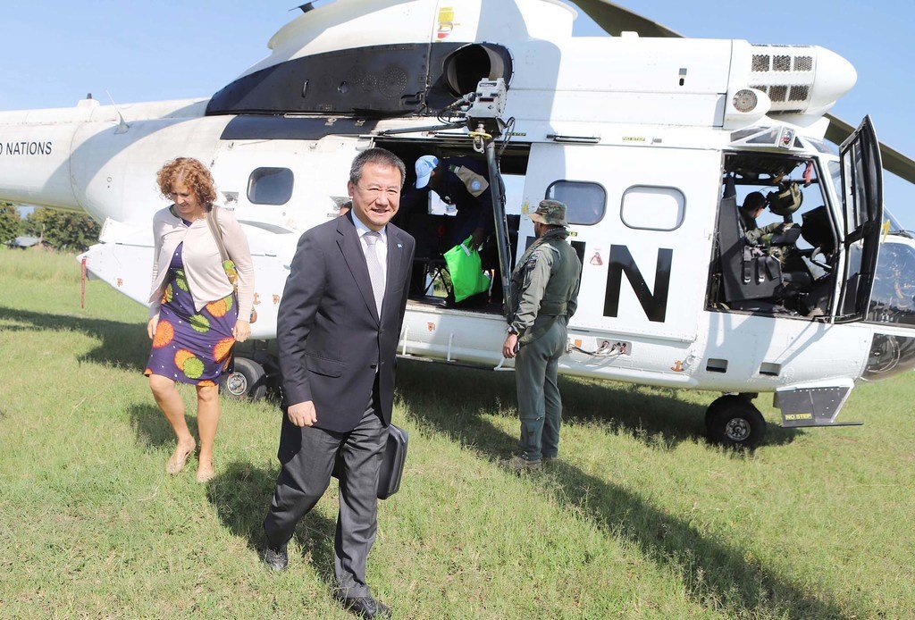 A man in a suit, identified as Mr. Huang Xia, walks on a grassy field away from a white United Nations helicopter. A woman in a colorful dress walks beside him.