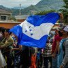 Un grupo de migrantes hondureños se encuentra en fila, con una persona ondeando prominentemente la bandera de Honduras. Están cerca de la frontera con Guatemala.