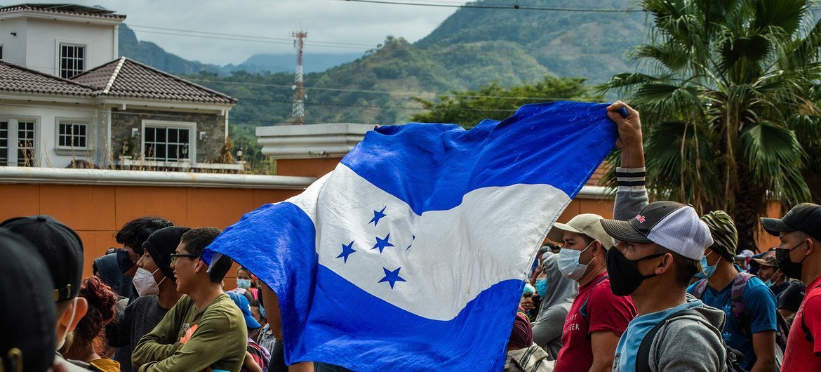 Un grupo de migrantes hondureños se encuentra en fila, con una persona ondeando prominentemente la bandera de Honduras. Están cerca de la frontera con Guatemala.