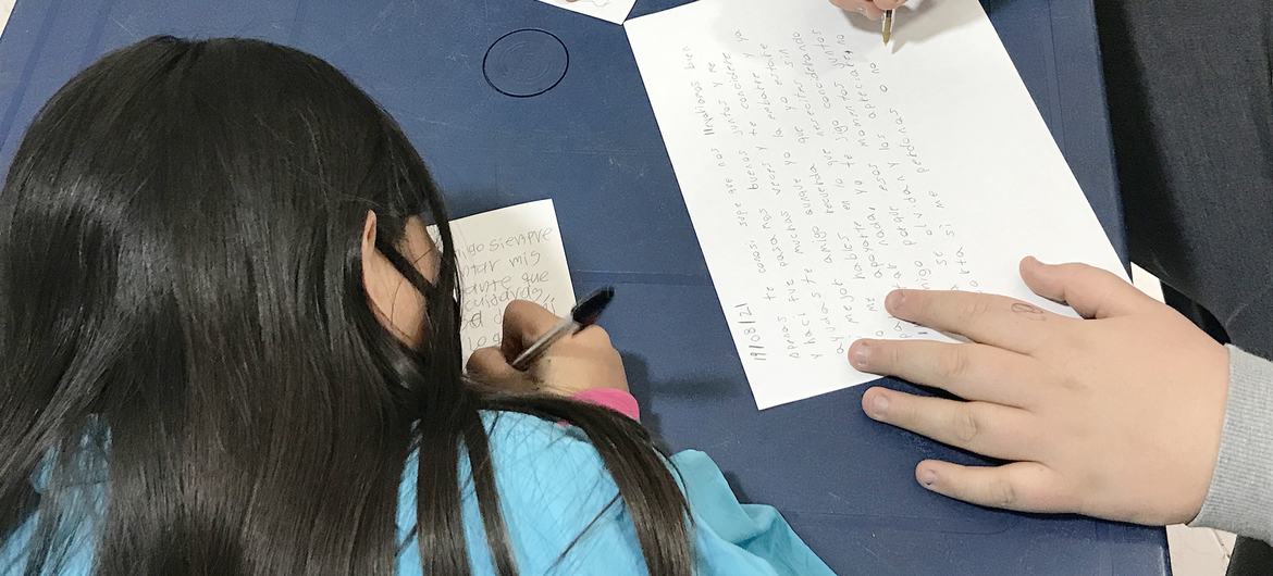 Crianças escrevem artigos de jornal sobre migração e semelhanças alimentares em uma mesa azul na Colômbia.