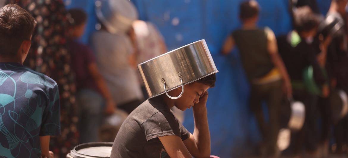Uma criança colocando uma panela na cabeça para se proteger do sol enquanto espera para conseguir comida Uma criança colocando uma panela na cabeça para se proteger do sol enquanto espera para conseguir comida
