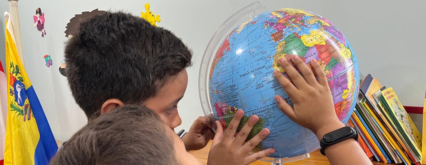 Dois meninos observam um globo terrestre sobre uma mesa, com a bandeira da Venezuela ao fundo.