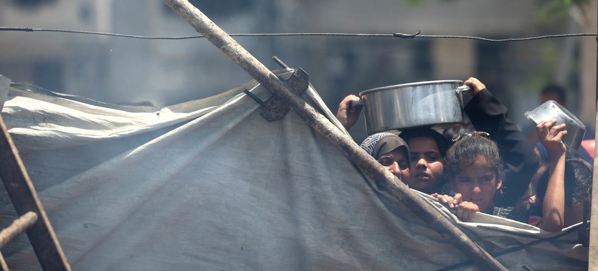 Tras una lona desgastada, niños de Gaza sostienen cuencos vacíos, con el rostro marcado por la fatiga y el hambre.