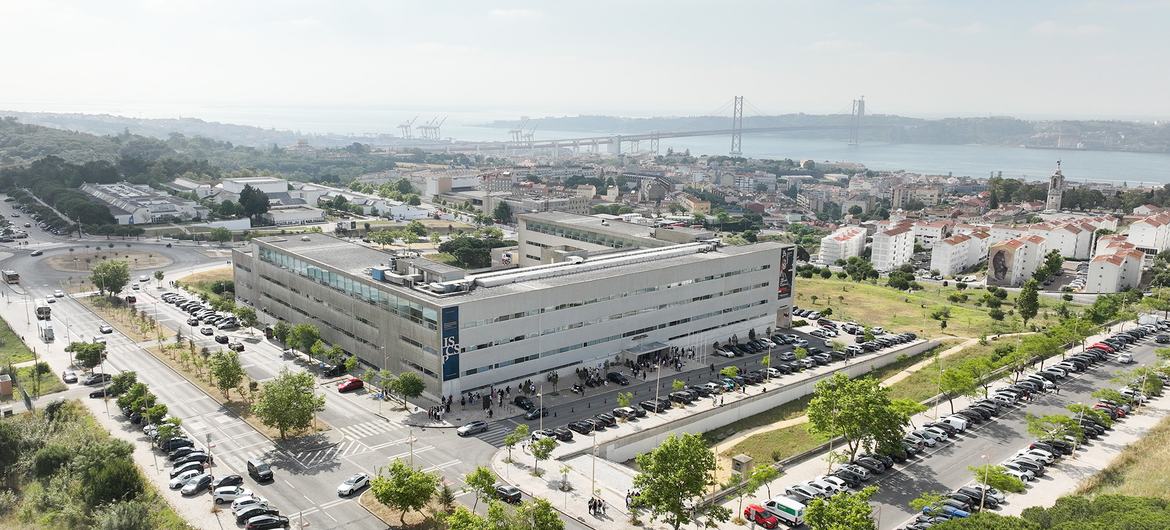 Vista aérea do campus do ISCTE-IUL em Lisboa, Portugal, com um edifício moderno e estacionamento, tendo a cidade e uma ponte como pano de fundo.
