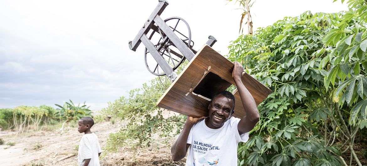 Um homem sorridente, vestindo uma camiseta branca, carrega uma máquina de costura de madeira no ombro, enquanto um menino caminha por perto em uma paisagem rural repleta de detritos.