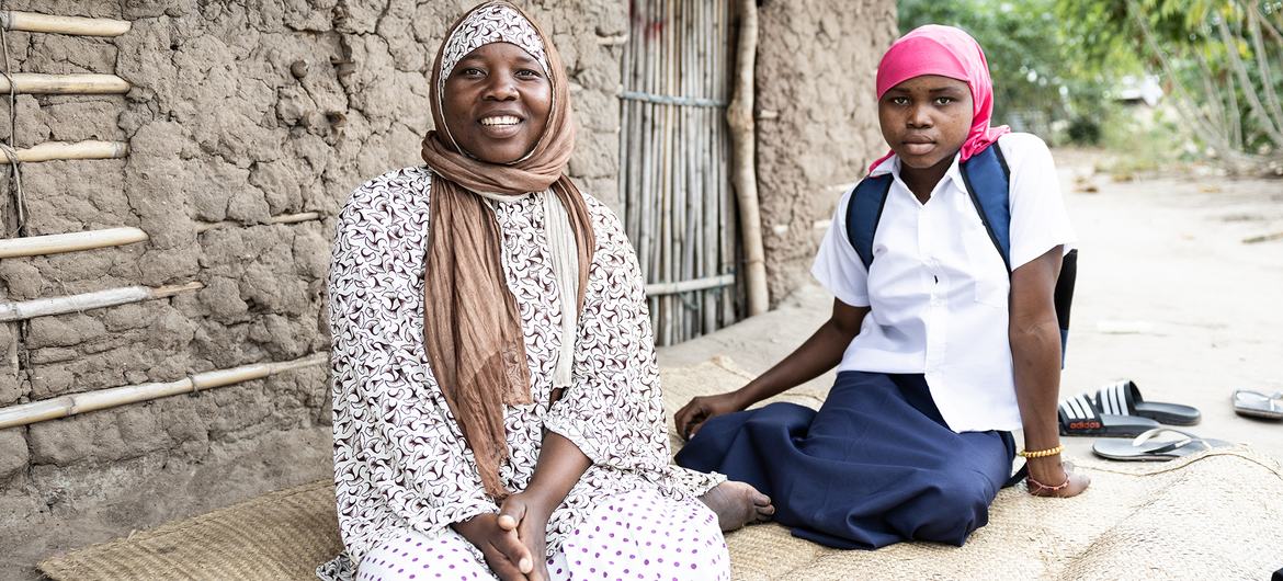 Uma mãe e sua filha sentam-se do lado de fora de uma estrutura de barro e bambu em Metuge, Moçambique, como parte de uma iniciativa da OIM para fornecer uniformes escolares a crianças deslocadas.