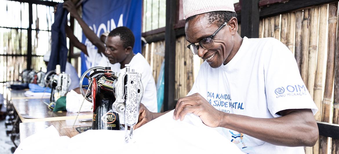 Um senhor africano de idade, usando óculos e boné, sorri enquanto trabalha em uma máquina de costura em uma oficina. Outros homens também estão presentes, trabalhando em máquinas de costura, ao fundo. O homem veste uma camiseta branca com a marca da OIM (Organização Internacional para as Migrações).