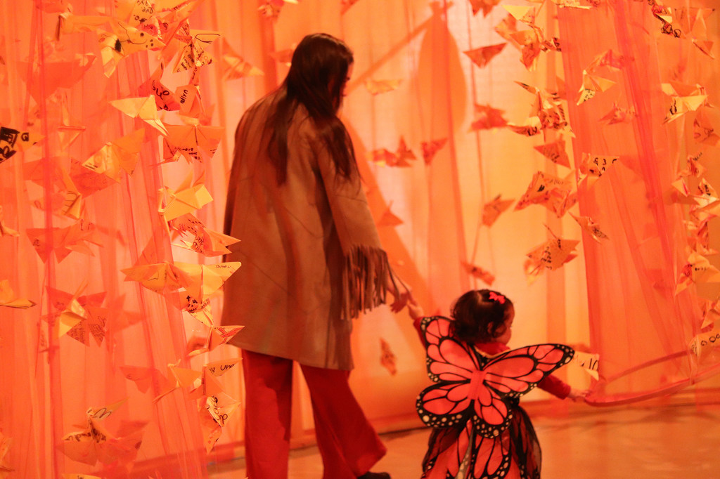 Una mujer y un niño pequeño disfrazado de mariposa caminan entre una instalación artística de grullas de origami naranjas colgantes.