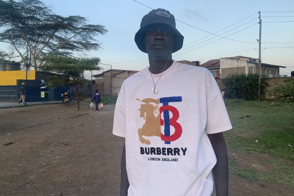 Picha ya kijana aliyevaa fulana nyeupe ya Burberry na kofia nyeusi ya ndoo, amesimama nje.