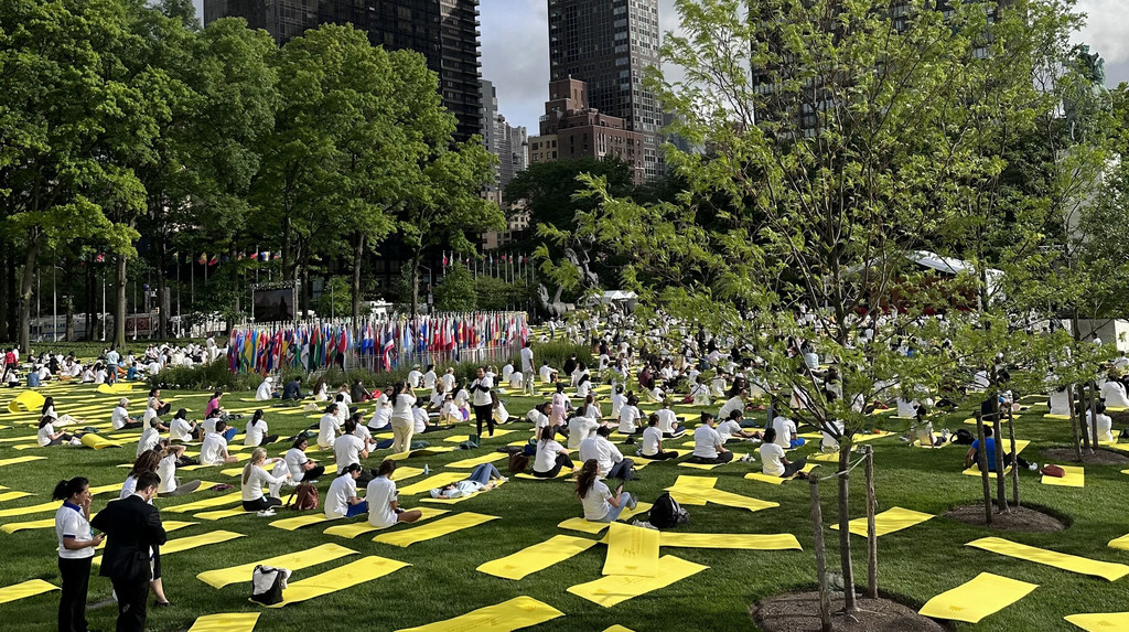 Un grand groupe de personnes participe à une séance de yoga sur une pelouse au siège des ONU à New York, avec de nombreux drapeaux internationaux en arrière-plan.