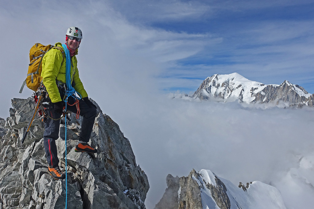 Um alpinista do sexo masculino está em um pico rochoso, usando capacete e equipamento de escalada, com montanhas nevadas e nuvens ao fundo.