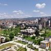 Vue panoramique de la ville de Pristina au Kosovo par une journée ensoleillée, montrant un mélange de bâtiments modernes et anciens, une mosquée imposante aux dômes blancs, un grand parc verdoyant et des montagnes en arrière-plan.