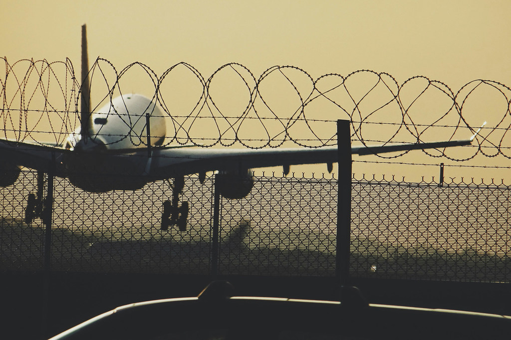 En el aeropuerto de Heathrow, en Longford, Reino Unido, se puede ver la silueta de un avión en una pista de aterrizaje tras una valla de tela metálica rematada con alambre de púas.
