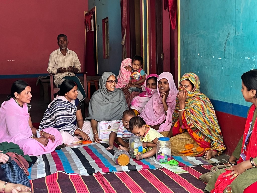 भारत के बिहार में एक सामुदायिक सत्र के दौरान, UNICEF की एक कार्यकर्ता रंगीन कालीन पर महिलाओं और बच्चों के एक समूह के साथ बैठी हुई एक दस्तावेज़ पकड़े हुए है।