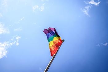 Un drapeau arc-en-ciel, symbole de la fierté LGBTQ+, flotte au vent sur un mât, se détachant sur un ciel bleu parsemé de quelques nuages.