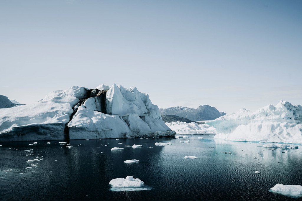 Icebergs e blocos de gelo flutuam em águas escuras sob um céu claro na paisagem da Groenlândia.
