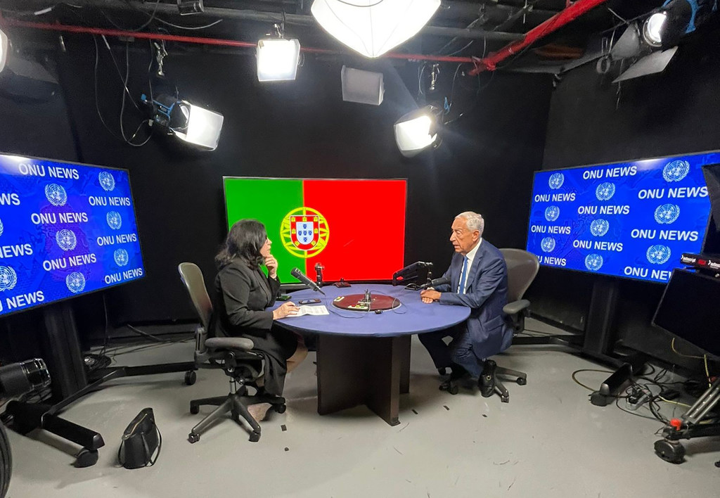 Marcelo Rebelo de Sousa é entrevistado por uma apresentadora de notícias em um estúdio da ONU News, com a bandeira de Portugal ao fundo.